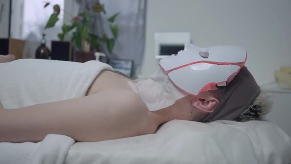 Side View of Relaxed Young Caucasian Man Lying on Beauty Bed in Spa Salon with LED Light Therapy