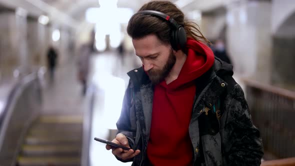 A Brutal Longhaired Man with a Beard Stands at a Metro Station and Listens to Music with Headphones