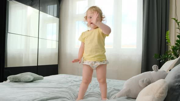 Funny Little Curly Blonde Girl Child in Bright Yellow Tshirt and White Shorts Stands on Bed During