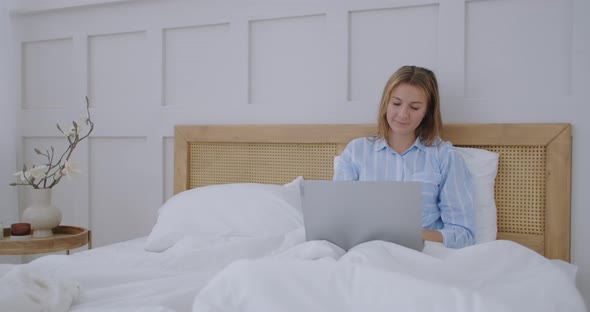 Young Millennial Girl Sitting on a Bed in the Morning, Uses Laptop Computer. Caucasian Woman Home