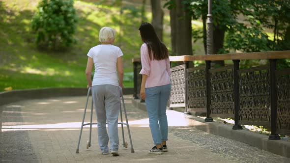 Volunteer Walking With Elderly Woman Using Walker in Hospital Park, Disability