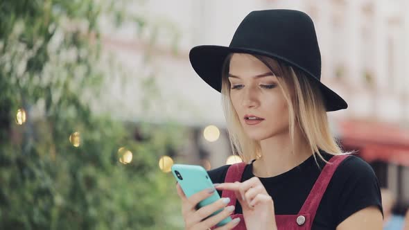 Pretty Young Blond Girl Wearing a Black Hat Scrolling on Her Smartphone and Standing Than Looking To