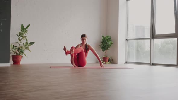 Young Woman in Red Sports Uniform Performs the Astavakrasana Asana in Yoga Studio