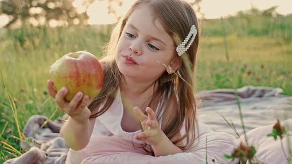 Little Cute Girl with Flowing Hair in a White Summer Dress Lying on a Green Lawn Eating an Apple