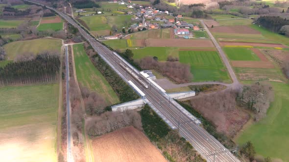 Aerial view of passenger train running through a lonely station past rural green fields. Tracking sh