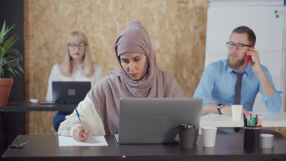 Muslim Woman Is Working in Office, Making Notes on Paper and Looking on Screen