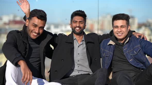 Three Happy Middle Eastern Men Waving Looking at Camera Smiling Sitting at Glass Fence on Rooftop