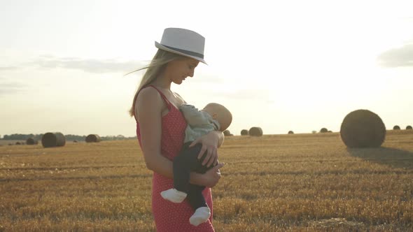 Pretty Young Mother Feeds Baby Son with Breast in Haystack Field in Light Day