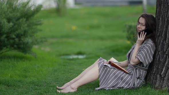 Happy Woman is Talking By Cell Phone in Park Sitting on Grass Under Tree at Summer Day