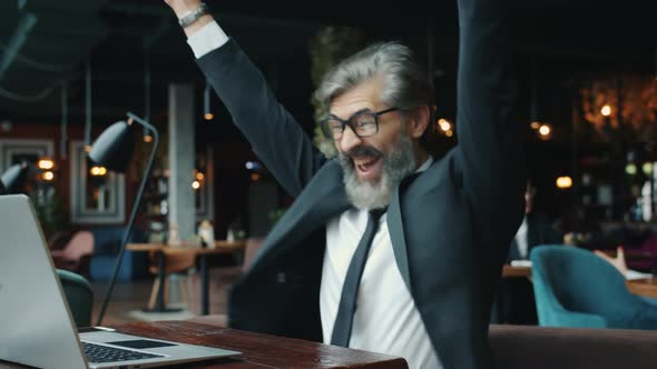 Excited Entrepreneur Mature Man Using Laptop in Cafe Then Raising Arms Celebrating Success