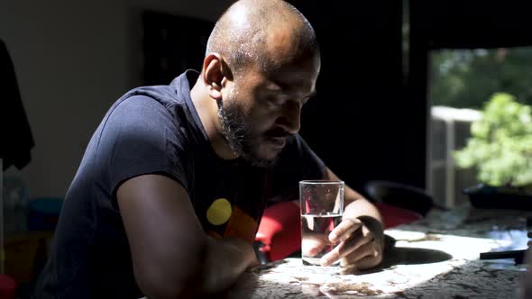 A casually dressed Indian man slumped over a counter table, the troubled man gazing into a glass of