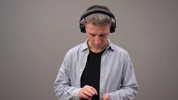 A Young Man on a Gray Background in Headphones Is Typing Text on a Smartphone