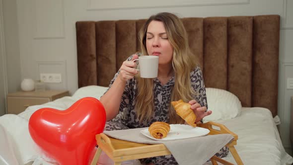 a Young Woman Sits in Bed and Has Breakfast on the Bedside Table Next to a Heart Balloon