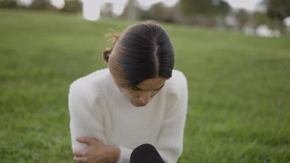 Worried Young Woman Sits on Grass Scratches Arm and Looks Down