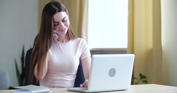 Portrait of Young Woman with Long Hair Sits at Home in Office at Work Table, Speaks on Phone To