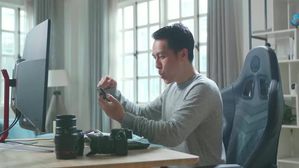 Happy Asian Cameraman Looking At Mobile Phone While Using Desktop Computer For Working At Home