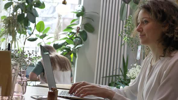 Positive Woman Drinks Coffee and Works on Laptop in Cafe