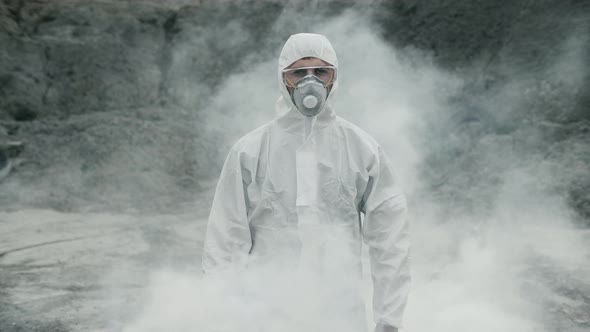 Lab technician in a mask and chemical protective suit, walks on dry ground through toxic smoke