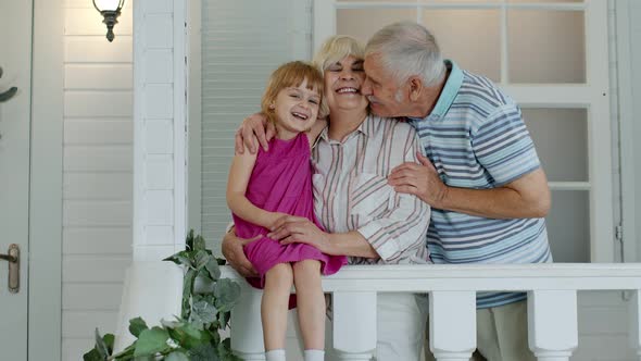 Senior Grandfather and Child Girl Embracing Grandmother and Making a Kiss in Porch at Home