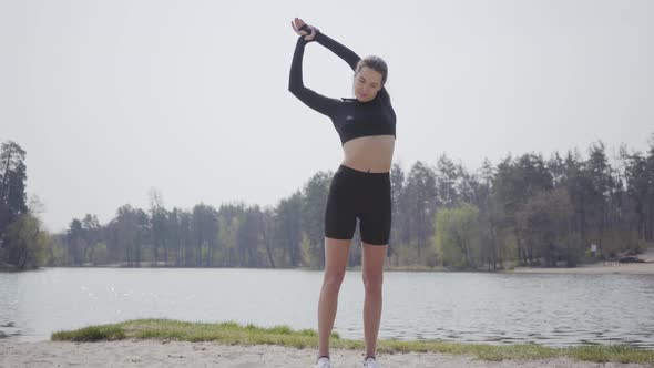 Charming Slim Young Woman Doing Sport Exercises, Standing on the Riverbank. Beautiful Landscape in