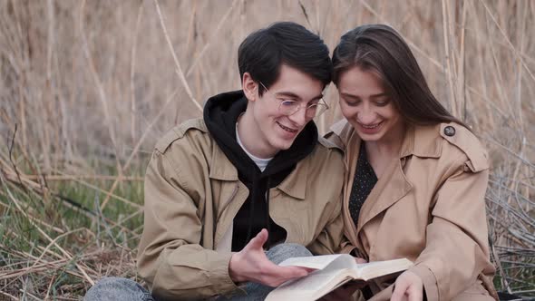 Young Couple Sitting in Field and Reading Book Aloud