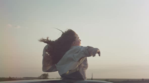 Young Beautiful Girl Rides in the Car with Raised Hands Up a Woman in the Sunroof of the Car