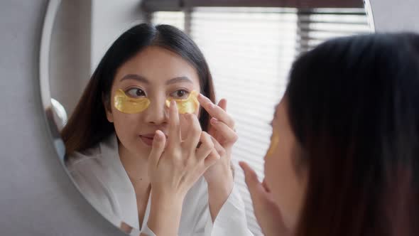 Beautiful Asian Lady Applying UnderEye Patches While Standing Near Mirror In Bathroom
