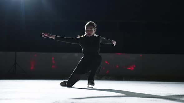Beautiful Young Sportswoman is Skating on Ice Rink in Darkness Performing Elements of Figure Skating