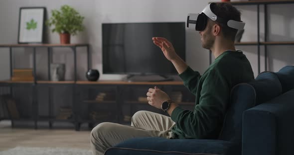 Adult Man Is Sitting on Couch in Living Room and Using Hmd Gadget with Virtual Reality Swiping