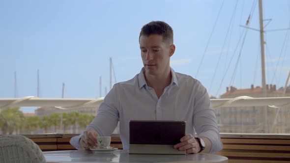 a young caucasian man at a café in a southern french harbour, using his tablet and drinking his coff