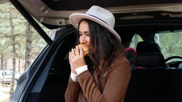 A young girl sits in the trunk of a car, eats a croissant, and drinks coffee. Woman in a hat