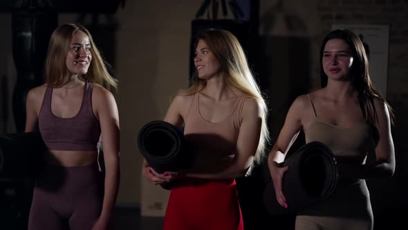 Three Smiling Fit Young Caucasian Women with Exercise Mats Walking in Dark Gym in Slow Motion