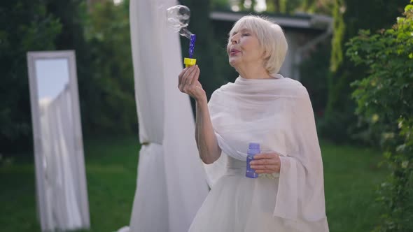 Carefree Caucasian Senior Woman in Wedding Dress Blowing Soap Bubbles in Slow Motion Standing