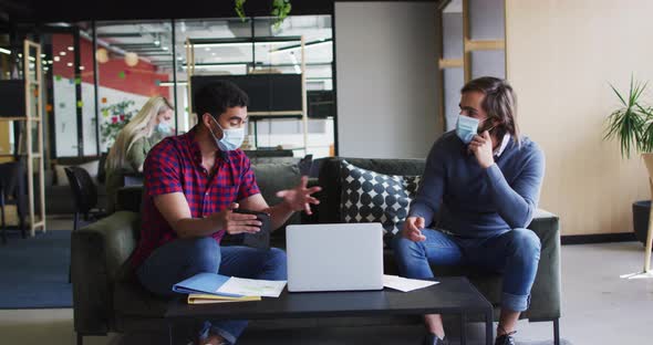 Diverse business people wearing masks using laptop and goign through paperwork in modern office