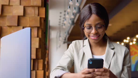 Young Indian Woman in Glasses Using Phone While Sitting in Cafe with Laptop