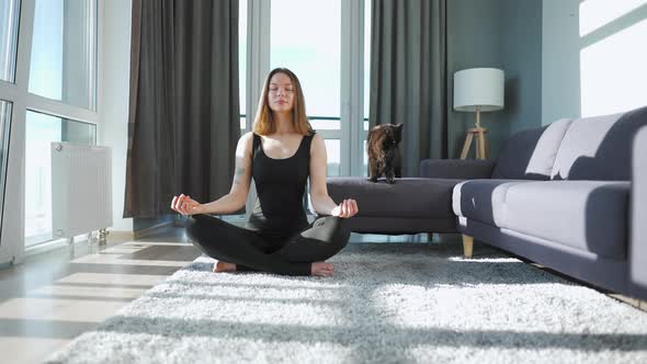 Young Caucasian Woman in Black Jumpsuit Sitting in Lotus Position and Meditating Yoga at Home