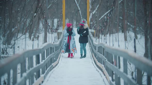 Two Young Women Walking and Having a Talk on the Snowy Bridge - Drinking Hot Drinks From the Thermos