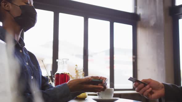 African american woman in face mask taking contactless card payment from customer at cafe bar