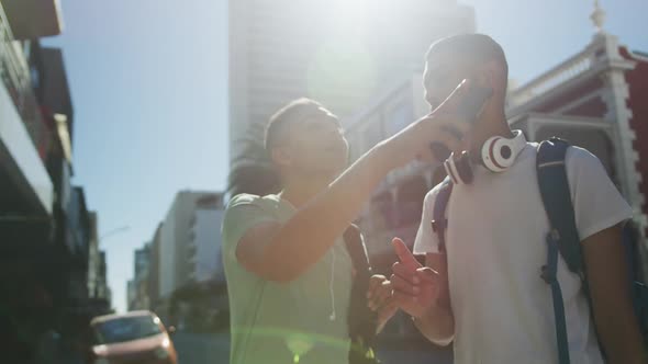 Two happy mixed race male friends standing, talking and using smartphone in the street