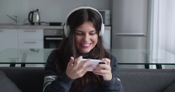 Millennial Brunette Girl in Headphone Play Game with Smart Phone on Modern Kitchen Background