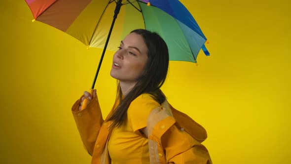 Cheerful Woman in a Yellow Raincoat with a Rainbow Umbrella is Checking for Rain