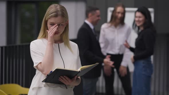 Stressed Sad Beautiful Young Woman Crying Standing Outdoors with Colleagues Mocking at Background