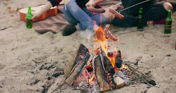 Close View of Friends Frying Sausages Sitting Around Bonfire Drinking Beer Playing Guitar on Sandy