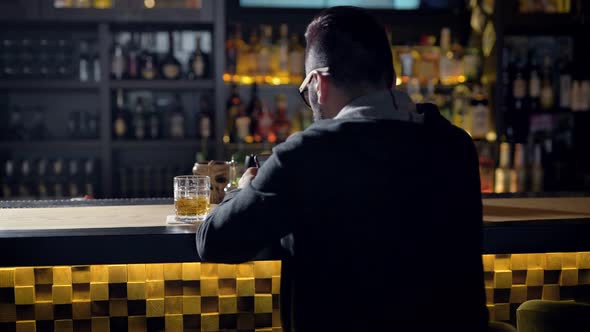 Behing Shot of a Man Quietly Sitting at the Bar with Glass of Whiskey, Chatting Online