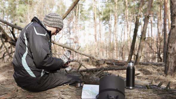 A tourist on a walk in a forest park drinks tea from a thermos and looks at a map on a smartphone.