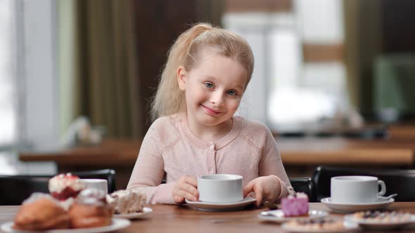 Happy Smiling Baby Girl Posing Holding Cup of Tea Sitting at Table