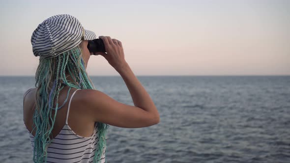 A Young Woman in a Marine Striped Dress Looks Through Binoculars at the Sea or Ocean