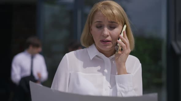 Confident Businesswoman Talking on Phone Standing Outdoors on Office Terrace with Blurred Colleagues