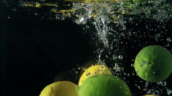 Close-up of falling ripe lemons and limes into sparkling water on black background, making cocktail