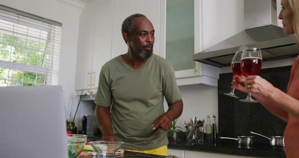 Diverse senior couple preparing food in kitchen using laptop and drinking wine